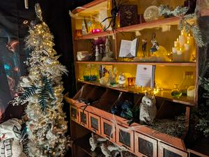 A display cabinet decorated for a magical or spooky holiday theme, featuring a white, 
                    lit Christmas tree on the left and shelves on the right filled with potion bottles, candles, spiderwebs, 
                    and an open book. A stuffed white owl sits near the lower drawers.