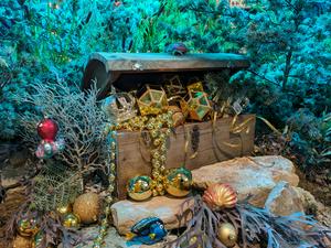 A treasure chest overflowing with gold ornaments and beads, set within a vibrant, 
                    teal-lit underwater-themed display featuring coral-like branches, fish, and shell decorations.