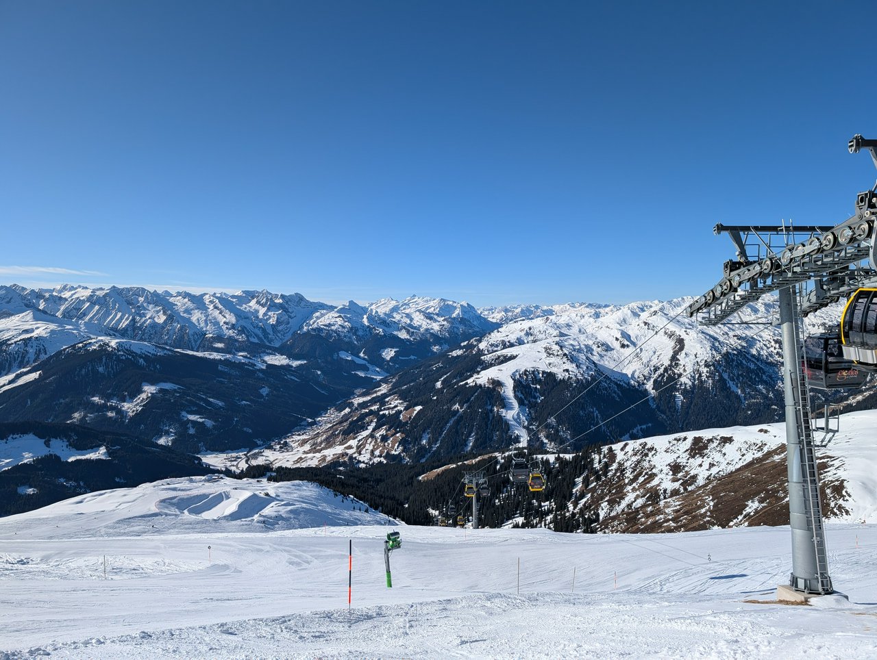 Standing on top of a prepared piste, a gondola lift on the right side, looking out at the mountains in the background. Clear blue sky, snow covered mountain tops, but still a lot of green pines, as there hasn't been fresh snow in a bit..