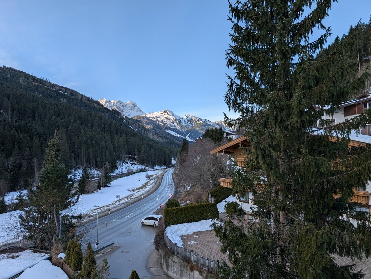 A winding street in a valley, a river runs next to it, pine trees cover the sides and in the distance a couple of snow covered mountain peaks.