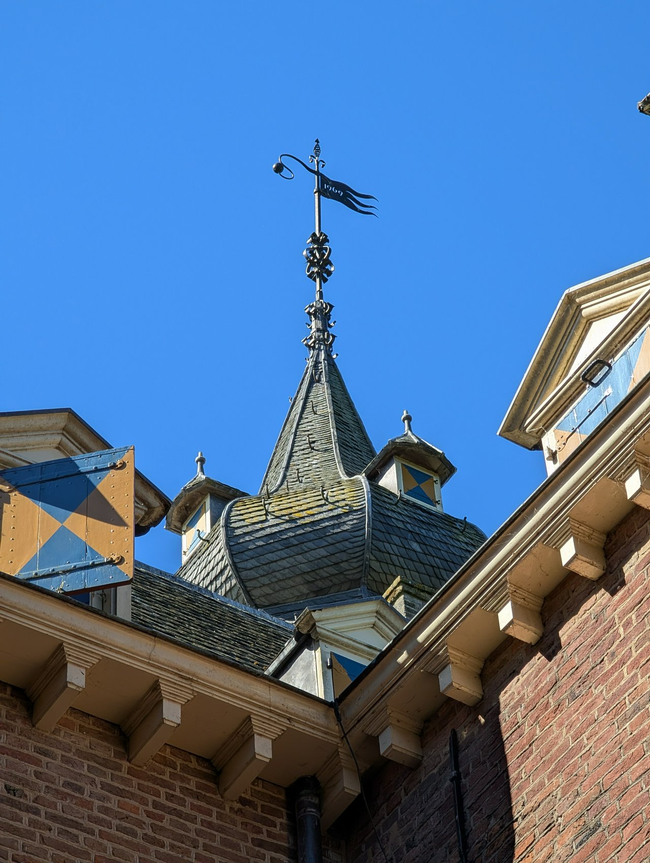Intricate roof, bell-shaped cupola, iron finial with a weather vane and blue and yellow heraldic shutters.